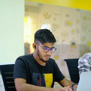 Young man with glasses using a laptop while wearing earphones in a modern office setting.