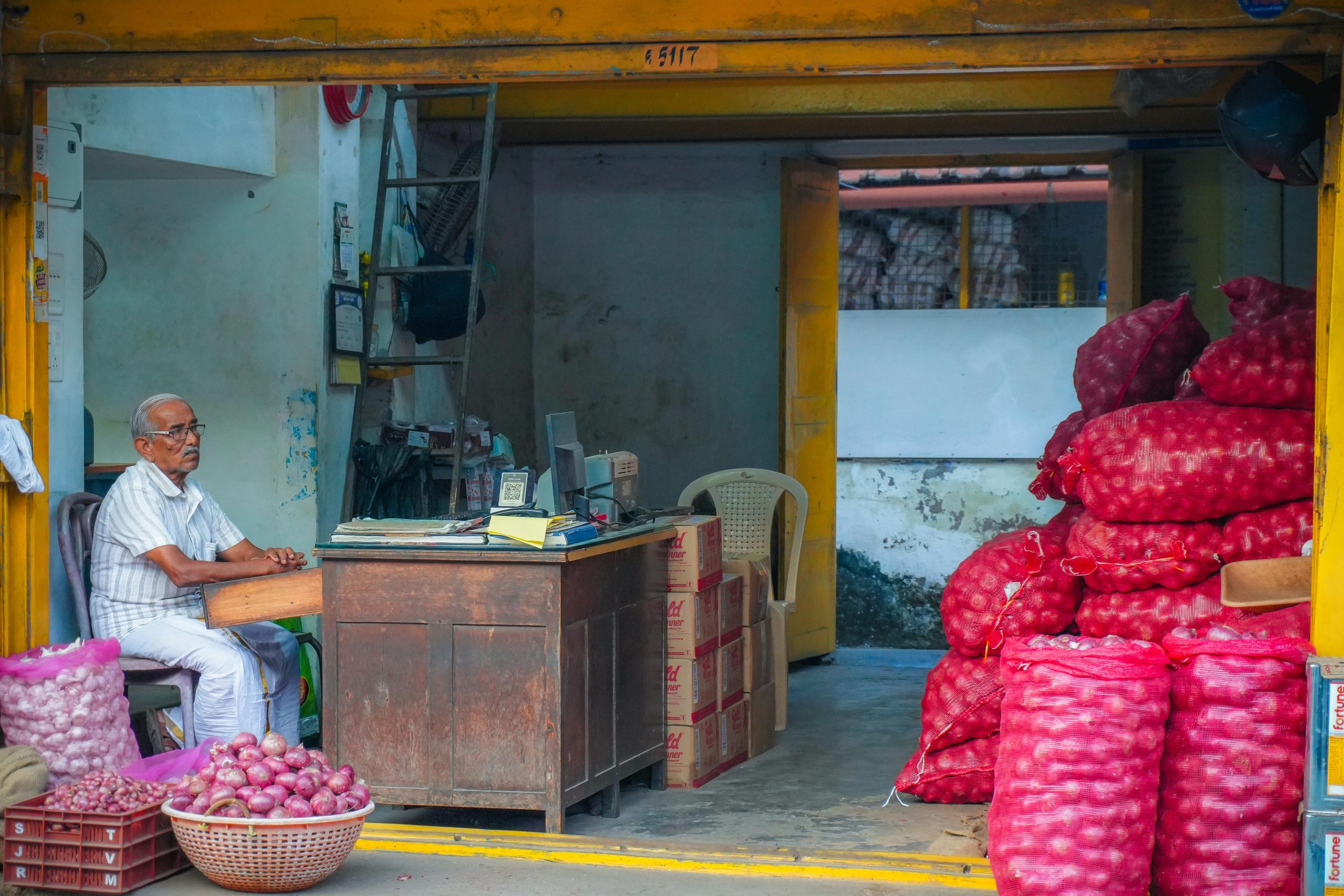 A man sitting in a small market shop surrounded by bags of onions and stacks of boxes, creating a vibrant scene.