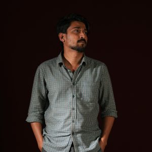 Contemplative man in checkered shirt stands against a dark background, hands in pockets.