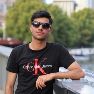 Portrait of a young man in sunglasses and t-shirt posing casually by a riverside in an urban setting.